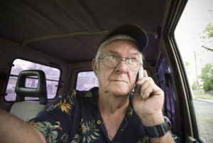 Older man in a tropical shirt talks on a cellphone inside a parked vehicle.