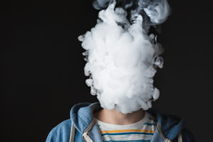 Person exhaling thick white vapor cloud, obscuring their face, against a dark background.