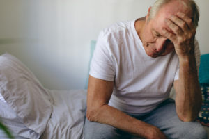 Older man sitting on a bed, holding his head with a distressed expression.