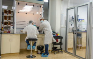 Two people in lab coats and masks work at a lab bench in a sterile laboratory setting.