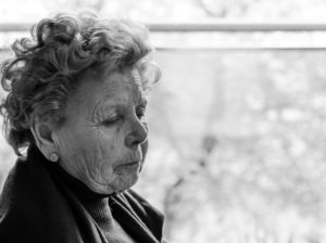 Elderly woman with curly hair sits in profile, eyes closed, in black and white.