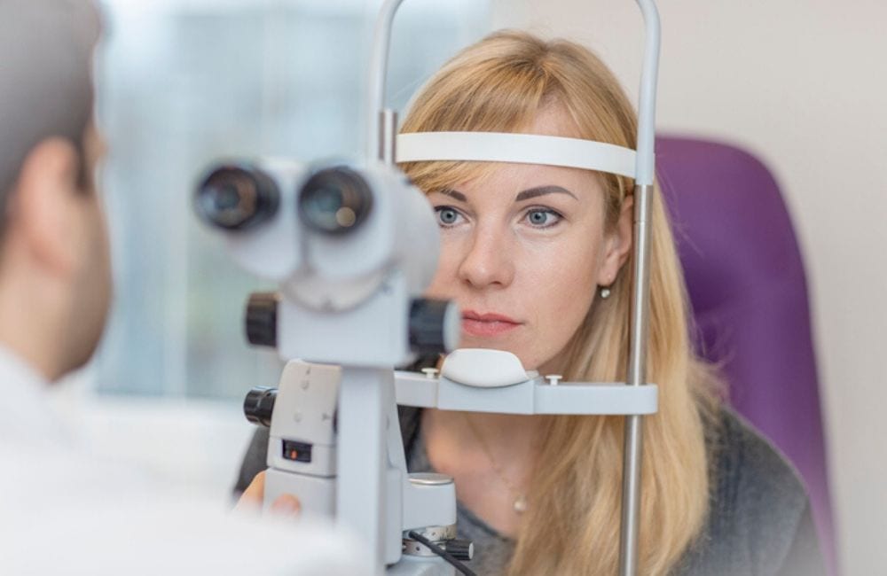 A red -haired woman in her late thirties receives an eye exam as she leans towards a phoropter. Elmiron's side effects may now include retinal maculopathy.