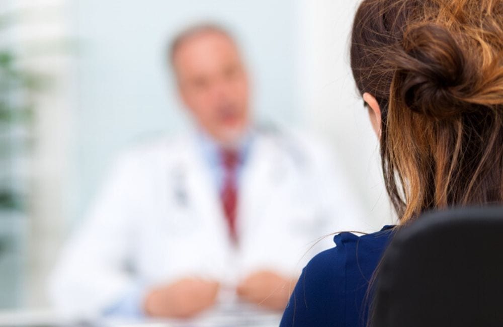 A woman is shown with her back to the camera, speaking to a doctor blurred out of focus. Women who use Elmiron are finding out about the risk of retinal maculopathy from their doctors, rather than Janssen Pharmaceuticals.