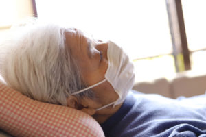 Elderly person with gray hair wearing a mask, lying on a pillow and resting.