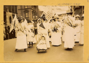 Women in white dresses march with flags and banners for "Votes for Women"; child in stroller.