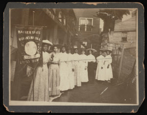 A group of women in white dresses stand in an alley, holding a "Banner State Woman's National" sign.