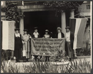 Six women stand outside holding a suffrage banner between two hanging striped flags.