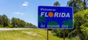 Blue "Welcome to Florida, The Sunshine State" sign by a roadside with trees in the background.
