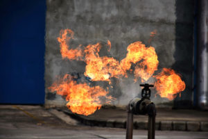 Flames emerge from a metal gas valve outdoors against a gray concrete wall.
