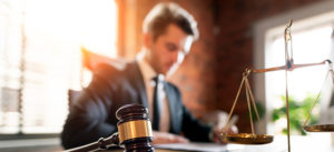 A gavel and scales of justice on a desk with a man in a suit working in the background.