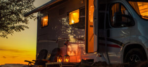 Camper van with open door and chairs outside at sunset, warm lights glowing inside.
