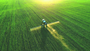 A tractor sprays crops in straight rows on a large green field, viewed from above.