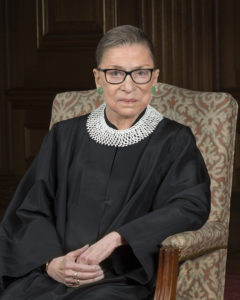 An older woman in judicial robes sits in a patterned armchair, looking at the camera.