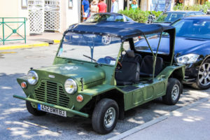 Green, open-top classic mini jeep parked on a street beside modern cars.