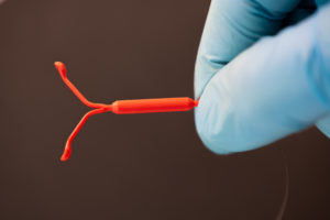 A gloved hand holds a red intrauterine device (IUD) against a dark background.