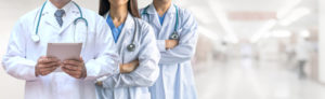 Three doctors in lab coats stand in a hospital hallway, two with arms crossed, one holding a tablet.