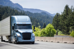 Blue big rig semi truck tractor with black grille transporting cargo in refrigerator semi trailer with reefer unit on the front wall running on the green forest highway road in Columbia Gorge