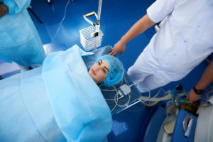 Patient lies on hospital bed in surgical gown, attended by medical staff in a clinical setting.