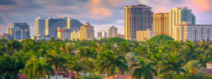 Skyline view of tall buildings and palm trees under a partly cloudy sky at sunset.