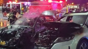 Two cars with severe fire damage; firefighters and emergency vehicles in the background at night.