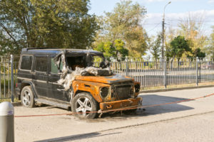 A severely damaged black SUV with a burnt front parked near a fence on a sunny day.