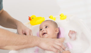 Baby in a bathtub with adult's hands nearby and rubber ducks on the tub edge.