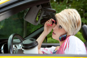 Woman in car applies mascara, holds phone with shoulder, and has coffee cup in foreground.