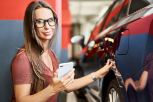 Woman holding a smartphone inspects the side of a parked car indoors.