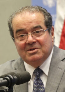 Man in a suit and tie speaking into a microphone, with a blurred flag in the background.