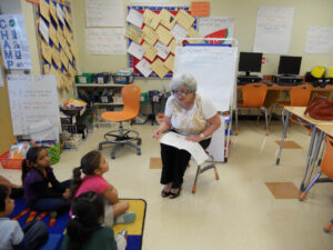 An older woman reads a book to a group of children seated on the classroom floor.